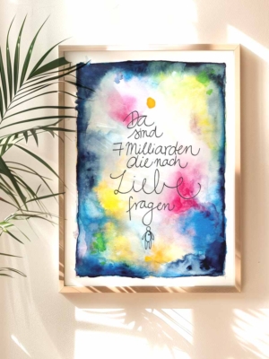 Ein gerahmter Kunstdruck mit einem künstlerischen Aquarellhintergrund in Blau, Gelb, Rosa und Grün. In der Mitte steht handgeschrieben: 'Da sind 7 Milliarden, die nach Liebe fragen'. Unter dem Text ist eine kleine, stilisierte Strichfigur. Ein Pflanze wirft einen Schatten auf Wand und Bild.