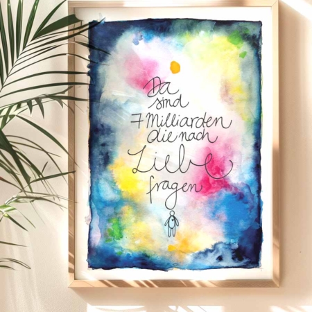 Ein gerahmter Kunstdruck mit einem künstlerischen Aquarellhintergrund in Blau, Gelb, Rosa und Grün. In der Mitte steht handgeschrieben: 'Da sind 7 Milliarden, die nach Liebe fragen'. Unter dem Text ist eine kleine, stilisierte Strichfigur. Ein Pflanze wirft einen Schatten auf Wand und Bild.