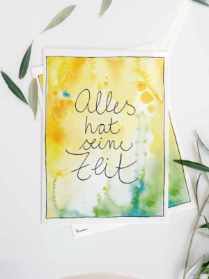 Eine Grußkarte mit Aquarellhintergrund in Goldgelb- und Grüntönen mit blauen Sprenklern. In der Mitte steht der Text „Alles hat seine Zeit".