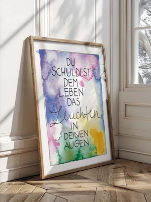 Eine farbenfroher Kunstdruck mit Aquarellhintergrund in Blau-, Lila-, Grün- und Gelbtönen. In der Mitte steht der Text „Du schuldest dem Leben das Leuchten in deinen Augen“ in einer Kombination aus Druck- und geschwungener Schreibschrift.