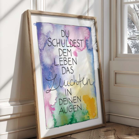 Eine farbenfroher Kunstdruck mit Aquarellhintergrund in Blau-, Lila-, Grün- und Gelbtönen. In der Mitte steht der Text „Du schuldest dem Leben das Leuchten in deinen Augen“ in einer Kombination aus Druck- und geschwungener Schreibschrift.