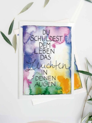 Eine Grußkarte mit Aquarellhintergrund in Blau-, Lila-, Grün- und Gelbtönen. In der Mitte steht der Text „Du schuldest dem Leben das Leuchten in deinen Augen“ in einer Kombination aus Druck- und geschwungener Schreibschrift. Die Karte liegt auf einem weißen Untergrund, dekoriert mit ein paar grünen Blättern.