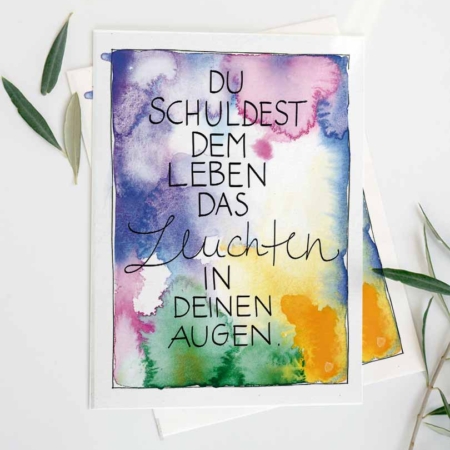 Eine Grußkarte mit Aquarellhintergrund in Blau-, Lila-, Grün- und Gelbtönen. In der Mitte steht der Text „Du schuldest dem Leben das Leuchten in deinen Augen“ in einer Kombination aus Druck- und geschwungener Schreibschrift. Die Karte liegt auf einem weißen Untergrund, dekoriert mit ein paar grünen Blättern.