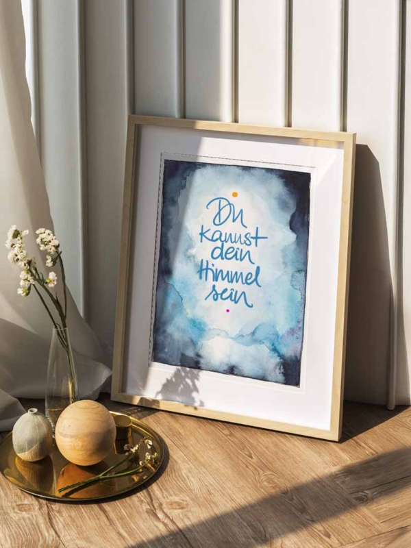 Ein gerahmtes Poster mit einem Aquarellhintergrund in Blau- und Weißtönen, das an Wolken oder Himmel erinnert. Im Zentrum steht in blauer Handschrift der Text „Du kannst dein Himmel sein“, ergänzt durch einen kleinen orangefarbenen Punkt oben und einen pinkfarbenen Punkt unten. Der Rahmen aus hellem Holz lehnt an einer weiß gestrichenen Wand, umgeben von natürlichem Licht. Im Vordergrund stehen auf einem goldenen Tablett eine Vase mit zarten weißen Blumen sowie zwei dekorative Kugeln aus Holz und Stein. Der Untergrund besteht aus hellem Holzfußboden.
