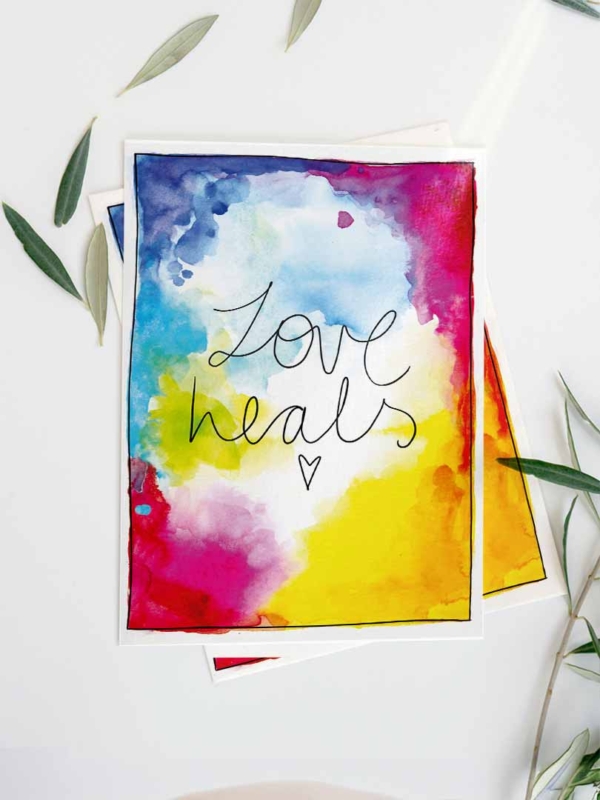 Eine Karte mit einem bunten Aquarell-Hintergrund in Blau-, Gelb-, Pink- und Rottönen. In der Mitte steht in schwarzer, handgeschriebener Schrift der Text 'Love heals.' Unter dem Text ist ein kleines, gezeichnetes Herz. Die Karte ist umgeben von grünen Olivenblättern.