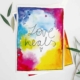 Eine Karte mit einem bunten Aquarell-Hintergrund in Blau-, Gelb-, Pink- und Rottönen. In der Mitte steht in schwarzer, handgeschriebener Schrift der Text 'Love heals.' Unter dem Text ist ein kleines, gezeichnetes Herz. Die Karte ist umgeben von grünen Olivenblättern.