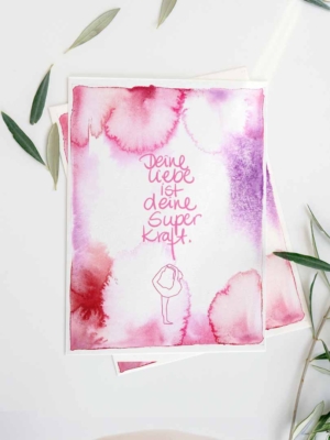 Eine Karte mit einem Aquarell-Hintergrund in Rosa- und Lilatönen. In der Mitte steht der handgeschriebene Text 'Deine Liebe ist deine Superkraft.' Darunter ist eine minimalistische Zeichnung einer stehenden Person in einer Yoga-Pose. Die Karte ist umgeben von grünen Olivenblättern.