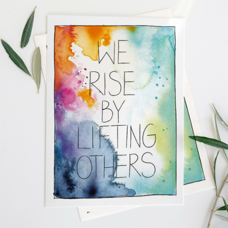 Eine Grußkarte mit Aquarellhintergrund in Orange-, Blau-, Grün- und Pinktönen. In der Mitte steht der Text „We rise by lifting others“ in Schreibschrift.