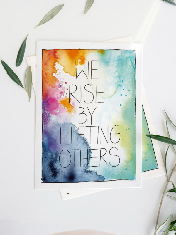 Eine Grußkarte mit Aquarellhintergrund in Orange-, Blau-, Grün- und Pinktönen. In der Mitte steht der Text „We rise by lifting others“ in Schreibschrift.