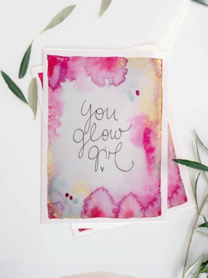 Eine Grußkarte mit Aquarellhintergrund in Gelb-, Gold- und Pinktönen. In der Mitte steht der Text „You Glow Girl“ in geschwungener Schreibschrift. Die Karte liegt auf einem weißen Untergrund, dekoriert mit ein paar grünen Blättern.