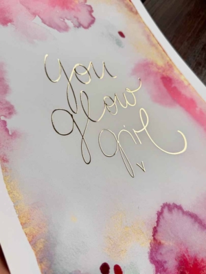 Perspektivische Aufnahme eines Aquarellkunstdruck in Gelb-, Gold- und Pinktönen. In der Mitte steht in Gold veredelt der Text „You Glow Girl“ in geschwungener Schreibschrift.