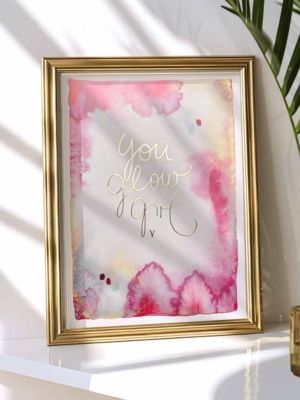 Ein gerahmter Kunstdruck mit Aquarellhintergrund in Gelb-, Gold- und Pinktönen. In der Mitte steht in Gold veredelt der Text „You Glow Girl“ in geschwungener Schreibschrift.