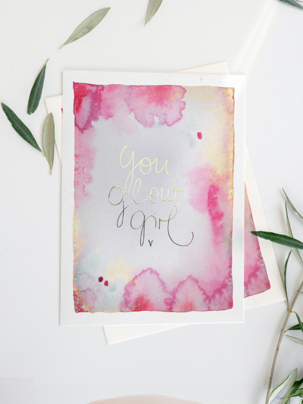 Eine Grußkarte mit Aquarellhintergrund in Gelb-, Gold- und Pinktönen. In der Mitte steht in Gold veredelt der Text „You Glow Girl“ in geschwungener Schreibschrift.