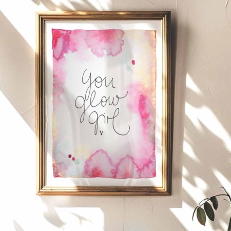 Ein gerahmter Kunstdruck mit Aquarellhintergrund in Gelb-, Gold- und Pinktönen. In der Mitte steht der Text „You Glow Girl“ in geschwungener Schreibschrift.