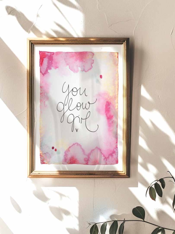 Ein gerahmter Kunstdruck mit Aquarellhintergrund in Gelb-, Gold- und Pinktönen. In der Mitte steht der Text „You Glow Girl“ in geschwungener Schreibschrift.